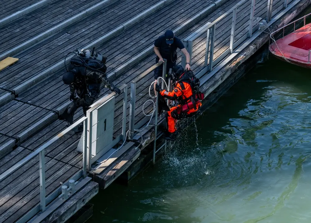 Plongeur de secours en tenue orange descendant dans l'eau depuis un ponton lors d'un exercice de sauvetage.